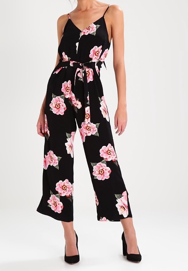 Combinaison noire à fleurs avec des fleurs roses, bretelles spaghetti, jambes larges et taille ceinturée. Portée avec des chaussures à talons noirs.