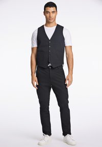 Zwarte vest met zes knopen aan de voorkant en twee steekzakken, gedragen over een wit t-shirt en gecombineerd met zwarte broek en witte sneakers.