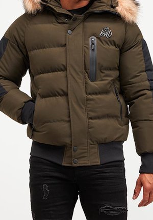 Homme portant une doudoune vert olive avec capuche bordée de fourrure, coudières noires, poche poitrine zippée et jean noir délavé.