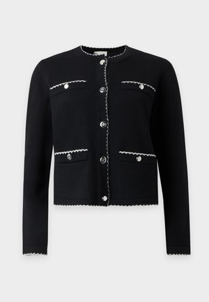 Femme portant une veste noire boutonnée avec des poches sur une jupe blanche à motifs arrivant aux genoux et des chaussures blanches pointues avec des nœuds.