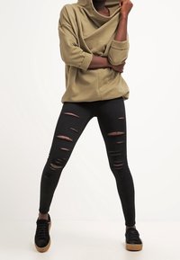 Sudadera con capucha en color oliva, de gran tamaño; jeans pitillo negros desgastados con roturas visibles; zapatillas negras con suelas marrones.