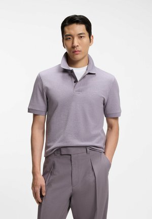 Hombre con polo color lila claro de cuello levantado y pantalones a juego, de pie con una mano en el bolsillo sobre fondo blanco.