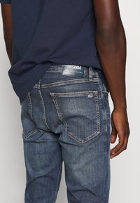 Mörkblå denimjeans med en figursydd design, med bakfickor och en märkesetikett på midjan. Texturen är slät med lätt blekning.
