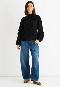Donna che indossa un maglione di maglia nero con motivi a cuore texturizzati, jeans a gamba larga blu e ballerine con stampa leopardata, in piedi davanti a uno sfondo bianco.