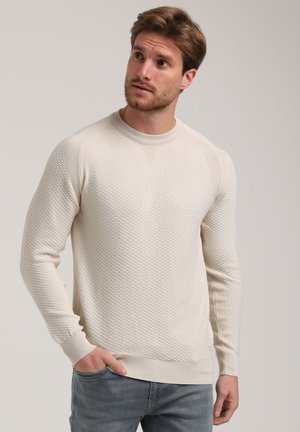 Light beige sweater with a textured, diamond-patterned knit. Ribbed neckline and cuffs, featuring a fitted design. Paired with light gray jeans.