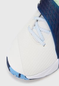 Valkoinen urheilukenkä, jossa on teksturoitu päällinen, siniset nauhat ja tummansinisiä yksityiskohtia. Ominaisuuksiin kuuluu tyylikäs muotoilu ja vahvistettu varvasalue.