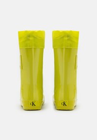 Botas de goma amarillas e impermeables con un acabado brillante, que presentan un puño de tela en la parte superior y un pequeño logo de cK en el talón.