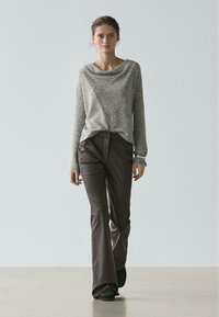 Jeune femme marchant, portant un pull gris texturé, un pantalon évasé marron foncé, des chaussures noires et un bracelet argenté, sur un fond clair uni.