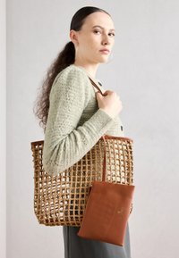 Geantă de umăr împletită de culoare bej cu detalii din piele maro, având un pouch detașabil din piele maro. Modelul poartă un pulover texturat de culoare verde deschis.