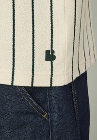 Tissu crème rayé avec des lignes verticales vertes, arborant une petite étiquette de logo verte ; associé à un denim noir avec des coutures jaunes.