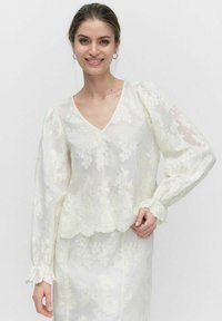 Hvid bluse med blomstret blonde-design, v-udskæring, puffede lange ærmer og bølget kant. Let og gennemsigtigt stof med teksturerede detaljer.