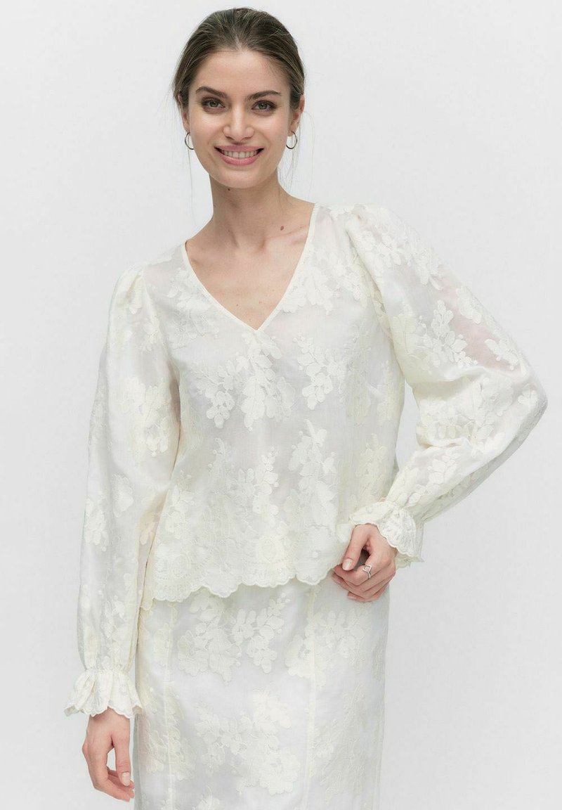 Hvid bluse med blomstret blonde-design, v-udskæring, puffede lange ærmer og bølget kant. Let og gennemsigtigt stof med teksturerede detaljer.