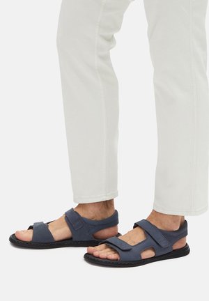 Niebieskie skórzane sandały z regulowanymi paskami i odkrytymi bokami, z teksturowanym wykończeniem oraz płaską czarną podeszwą. Noszone z białymi spodniami.