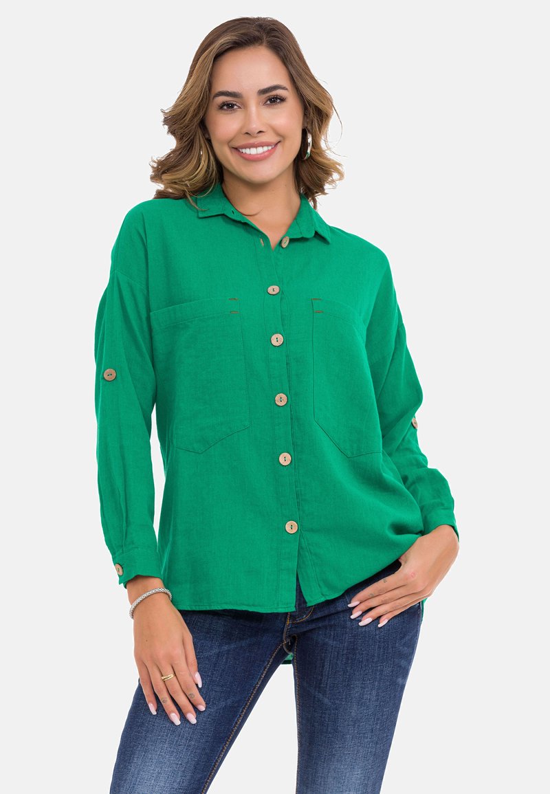 Groene linnen blouse met lange mouwen, twee borstzakken en houten knopen. Gladde textuur en ontspannen pasvorm, gecombineerd met donkere jeans.