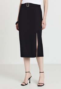 Schwarzer Midirock mit einem Frontschlitz, strukturiertem Material und einem passenden Gürtel mit quadratischer Schnalle. Getragen mit schwarzen Riemchenschuhen.