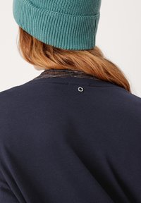 Maglione navy con scollatura rotonda, texture a coste e un dettaglio metallico vicino al collo sul retro. È visibile anche un berretto in maglia verde petrolio.