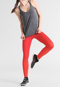Szary sportowy top na ramiączkach, połączony z jasnoczerwonymi legginsami z teksturowanymi wzorami. Czarne buty do biegania z różowymi akcentami.
