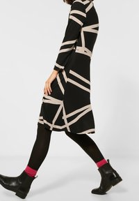 Musta mekko beigen abstraktien raitojen kanssa, pitkät hihat ja levenevä helma. Yhdistetty mustiin nilkkasaappaisiin ja vaaleanpunaisiin sukkiin, joissa on sileä kangas.