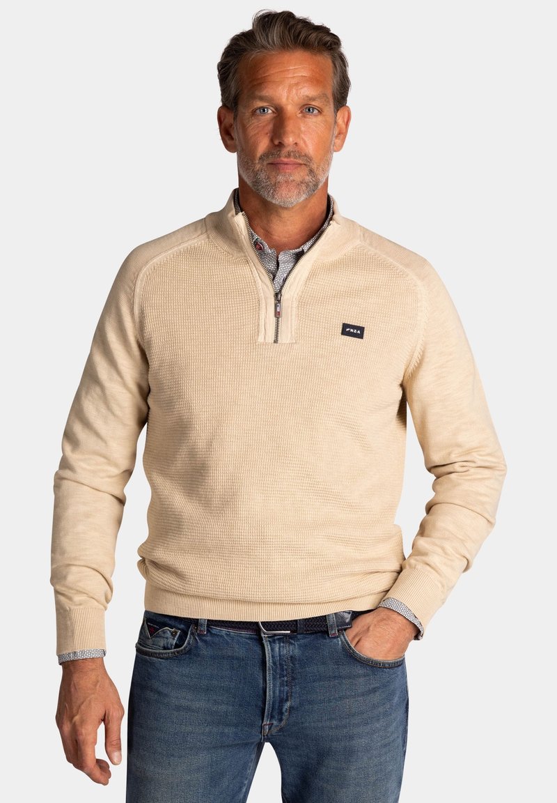 Beige ritssluitingen trui met geribbelde textuur en een klein logo op de borst, gecombineerd met blauwe jeans met een donkere riem en een ruitpatroon.