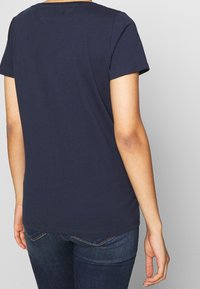 Person som bär en enfärgad marinblå t-shirt med kort ärm och mörkblå jeans, visad bakifrån mot en ljusgrå bakgrund.