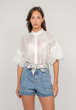 Femme portant un chemisier blanc transparent à manches bouffantes, un short en denim taille haute et une ceinture en tissu à motif nouée en nœud.