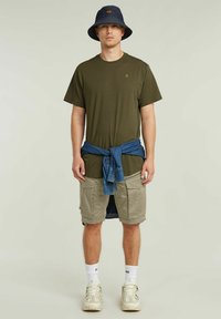 T-shirt verde oliva, pantaloni cargo color beige chiaro con tasche, camicia di jeans annodata in vita, cappello a secchiello blu navy e sneakers spesse.