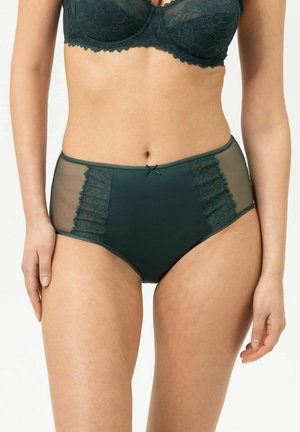 Culotte taille haute vert foncé avec dentelle et panneaux latéraux transparents, portée par une femme debout devant un fond uni.