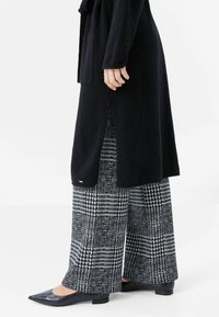 Svart lång kappa med slips, med en sidoklyfta. Under kappan, byxor i rutmönster i svart och vitt, i bred passform, tillsammans med spetsiga svarta skor.