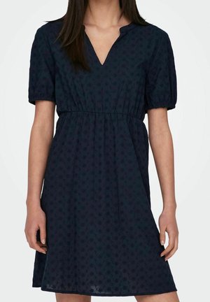 Kvinde iført en kortærmet kjole i mørk marineblå med subtile blomsterprint og en opsamlet talje, stående med afslappede arme.