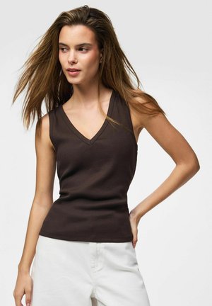 Jeune femme aux longs cheveux bruns portant un haut marron foncé sans manches à col en V et un pantalon blanc, posant avec une main sur la hanche.