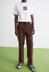 Bílý tričko s krátkým rukávem a hnědým logem, v kombinaci s hnědými kordovými kalhotami. Oblečené s šedými a černými teniskami, stojící na zelené podlaze.