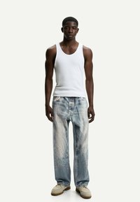 Canotta bianca senza maniche, jeans strappati leggeri blu con decorazioni, e scarpe beige. Texture liscia e vestibilità rilassata.