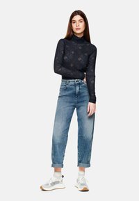 Schwarzes Spitzen-Turtleneck-Oberteil mit geometrischen Mustern, kombiniert mit hellblauen, hochgeschnittenen Jeans und grauen Sportschuhen mit weißen Akzenten.