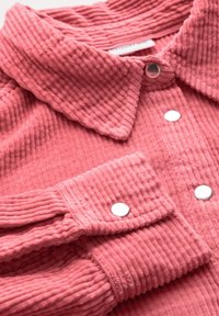 Camisa de pana en rosa suave con una superficie texturada y acanalada. Presenta un cuello con solapa y botones a presión plateados en la abertura y los puños.
