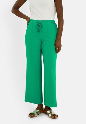Persona que lleva pantalones de pierna ancha de color verde brillante con cintura de cordón, top blanco sin mangas, anillos dorados y sandalias metálicas tipo slide.