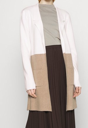 Personne portant un long cardigan blanc et beige avec blocs de couleurs, un haut gris et une jupe plissée marron foncé, debout devant un fond uni.