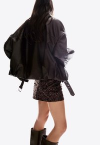 Zwarte leren oversized jas met glanzende textuur. Korte, bruin, gepailleerde rok met glanzende accenten, in combinatie met zwarte laarzen tot aan de knie.