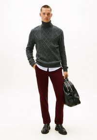 Grijze kabelgebreide coltrui, bordeaux corduroy broek, zwarte leren tas en zwarte instapschoenen. Model staat tegen een witte achtergrond.