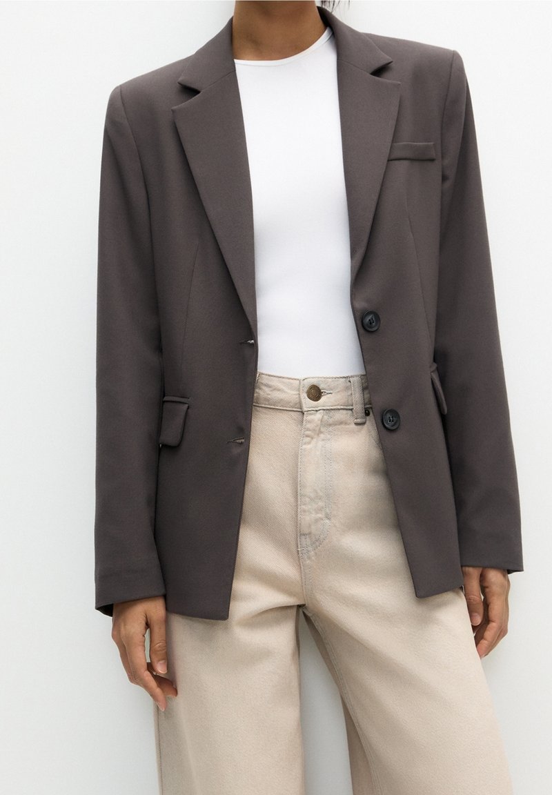 Blazer gris foncé avec des revers structurés, deux boutons et des rabats de poches avant, associé à un pantalon taille haute beige clair et un haut blanc.