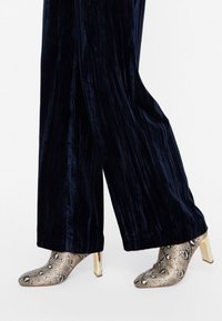 Stivaletti in pelle di serpente con tacco oro metallico, abbinati a pantaloni wide-leg in velluto scuro e morbido con strisce verticali.