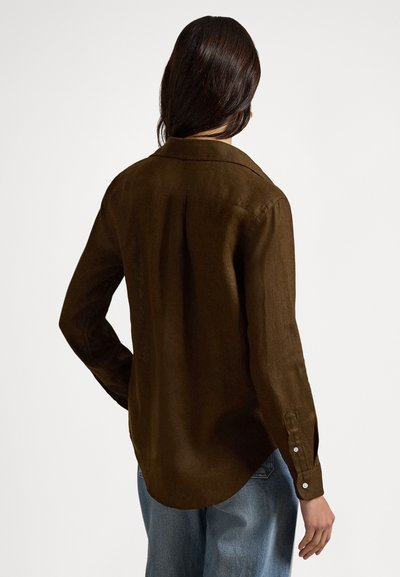 Brūns ar garām piedurknēm krekls no fakturēta auduma ar apkakli, noapaļu apakšu un pogu aprocēm, kopā ar gaiši zilām džinsa biksēm.