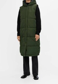 Grüne Steppweste mit Kapuze, mit einem gesteppten Design, Druckknöpfen und einem langen Schnitt, kombiniert mit schwarzen Stricksachen und einer Hose.