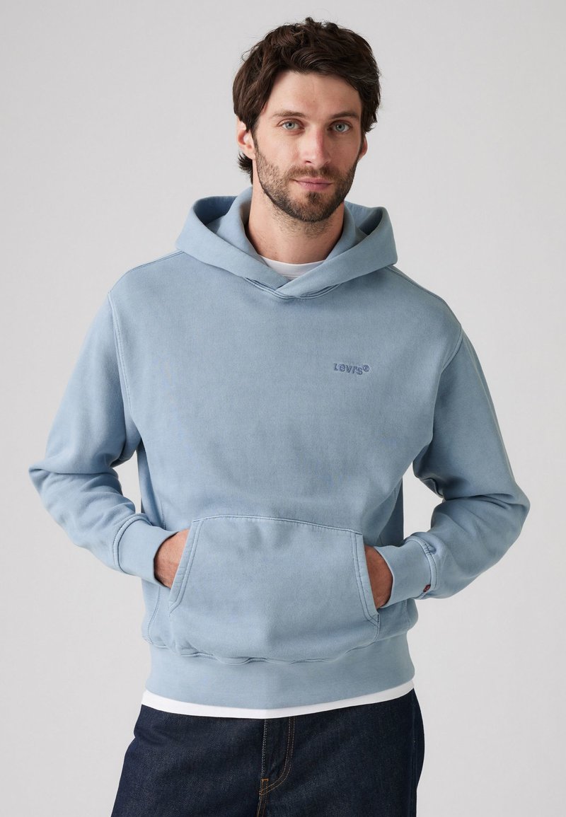 Lys blå hettegenser med kengurulomme, ribbestrikkede mansjetter og hem. Har en liten brodert logo på brystet, laget av mykt stoff.