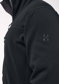 Giacca softshell nera con chiusura con zip, colletto alto e logo sulla spalla sinistra. Il materiale sembra liscio con una leggera texture.