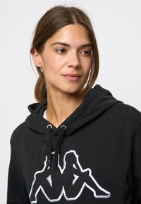Zwarte hoodie met een kap, koordaccenten en een opvallende witte geborduurde graphic. Zachte stof met een ontspannen pasvorm.