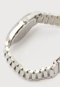 Πλάγια όψη ασημένιου μεταλλικού ρολογιού χειρός με συνδεδεμένο βραχιόλι και κυρτό κουμπί στην κάσα, πάνω σε απλό φόντο.