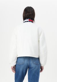 Biela skrátená bunda s dlhými rukávmi, s námorníckym golierom s červenými a bielymi pruhmi a logom "TOMMY JEANS". Džínsy sú viditeľné nižšie.