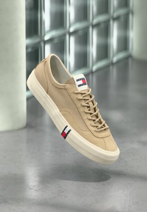 Beežid lõuendist spordijalatsid valge tallaga, paeltega ja Tommy Jeans logo keelega ning küljel, eksponeeritud klaasplokkide seina taustal.