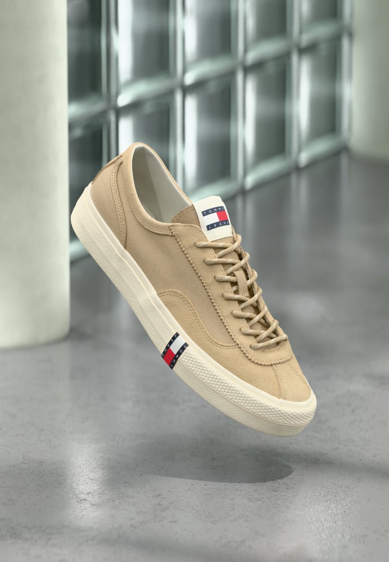 Μπεζ πάνινο αθλητικό παπούτσι με λευκή σόλα, κορδόνια και λογότυπο Tommy Jeans στη γλώσσα και στην πλευρά, παρουσιάζεται μπροστά σε φόντο τοίχου από γυάλινα τούβλα.