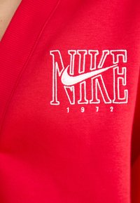 Red cotton sweatshirt features large white embroidered "NIKE" logo with a swoosh and "1972" detail at the bottom. Smooth texture and ribbed sleeves.